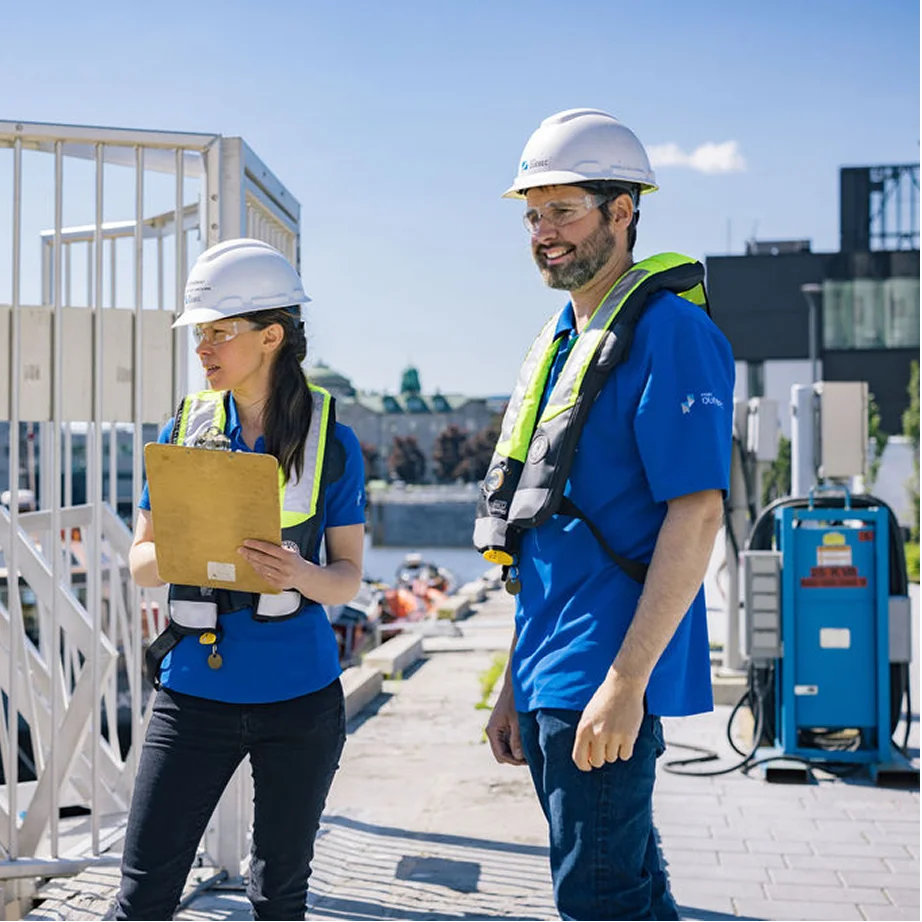 Deux travailleurs en casque et gilet de sécurité inspectant un chantier avec un clipboard près de l’eau.